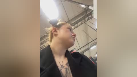 Media: Video of a fair-skinned woman with blonde hair in a ponytail, wearing a black jacket, with a tattoo of deer antlers on her chest. Background shows an industrial warehouse with exposed beams and fluorescent lighting.