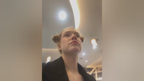 Media: Video of a young woman with blonde hair styled in two buns, wearing a black jacket, standing indoors under a curved, ceiling-mounted light fixture.