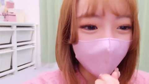 Media: Video of a young woman with long, light brown hair, wearing a pink surgical mask, standing in a white room with a green curtain and white shelves.