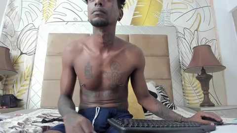 Media: Video of a shirtless, muscular, light-skinned Black man with tattoos, sitting at a desk in a bedroom with zebra-print curtains and a beige headboard.