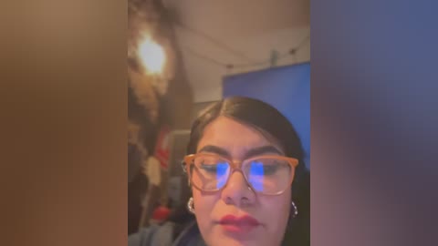 Video of a young woman with medium skin tone and long black hair, wearing large glasses and red lipstick, against a dimly lit, cluttered room with a hanging light and indistinct background objects.