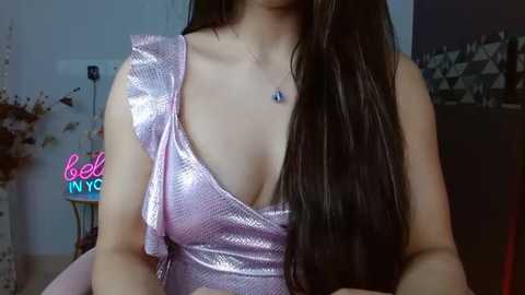 Media: Video of a woman with long, dark hair wearing a glittery pink one-shoulder dress, showcasing her cleavage and a necklace. Background shows a partially visible room with a neon sign and a dark wall.