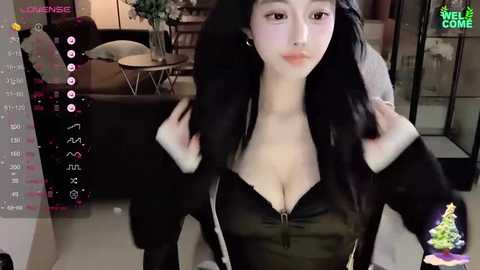 Media: Video of a young Asian woman with long black hair, wearing a black, low-cut, zipped-up top, adjusting her fur-trimmed jacket, in a modern, dimly-lit living room with a couch, plants, and a colorful Christmas tree in the background.