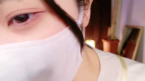 Media: Close-up video of an Asian woman with light skin, wearing a white surgical mask partially covering her mouth, with her brown hair tied back. Background features a potted plant and a mirror.