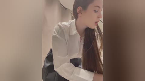 Media: A video of a young woman with fair skin, long brown hair, and a white shirt, leaning against a beige wall, wearing a black handbag.