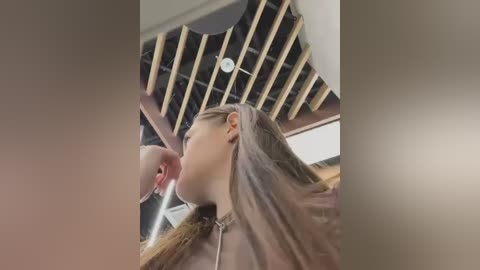 Media: Video of a young woman with long, brown hair in a beige jacket, performing oral sex on a man with a shaved head, set in a modern room with exposed wooden ceiling beams and a circular light fixture.