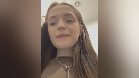 Media: Video of a young Caucasian woman with long, straight brown hair, fair skin, and blue eyes. She wears a beige top and a silver necklace with a heart pendant. The background is blurry, likely an indoor setting with soft lighting.