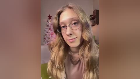 Media: Video of a young woman with long, wavy blonde hair and fair skin, wearing glasses, a pink top, and a necklace. Background shows a Christmas tree with pink and white decorations.