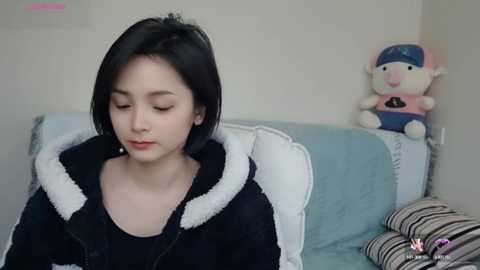 Media: Video of a young Asian woman with short black hair, wearing a black hoodie with white fur trim, sitting on a couch. Background features a stuffed toy and striped pillows, in a cozy, well-lit room.