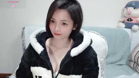 Media: Video of a young East Asian woman with straight black hair, wearing a black and white fuzzy robe, sitting on a light blue couch with plush toys.