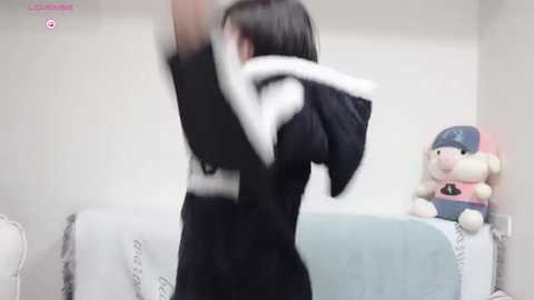 Media: Video of a woman in a black robe with white trim, holding her arms up, blurred, in a white room with a blue towel, plush toys, and a pink stuffed animal.