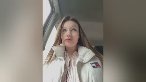 Media: A video of a young woman with long, straight brown hair, wearing a white puffy jacket with a red and black patch, looking out a car window with a neutral expression.