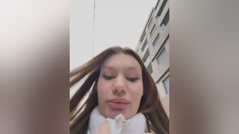 Media: A video of a young woman with straight, light brown hair, fair skin, and a white shirt, looking down and smiling. She is standing in front of a modern building with balconies, taken from a low angle.