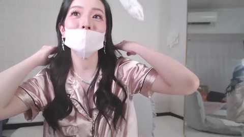 Media: Video of an Asian woman with long black hair, wearing a silver satin blouse and white surgical mask, adjusting a white feathered headpiece in a modern, white-walled room.