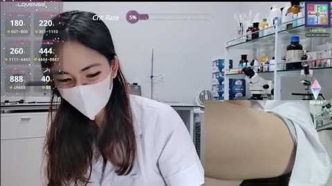 Video of an Asian woman in a white lab coat and mask examining a patient's leg in a clinical setting. Background shows medical equipment and supplies.