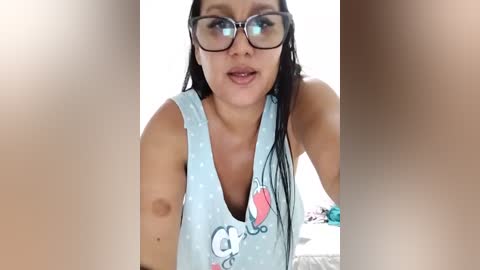 Media: Video of a woman with long dark hair, wearing large black-framed glasses, a light blue polka-dotted tank top, and speaking with parted lips. Background is blurred, possibly indoors.