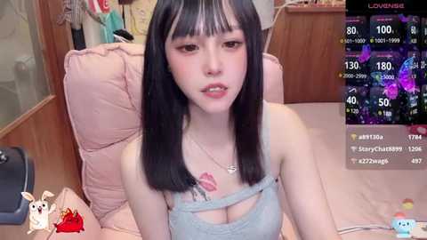 Media: A video of an East Asian woman with long black hair, light skin, and a slender physique, wearing a light blue spaghetti-strap top, sitting on a pink plush chair in a cozy room with a wooden background.