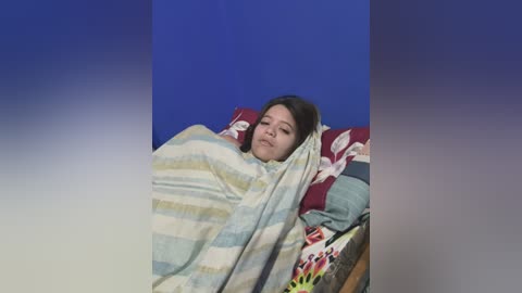 Media: Video of a young woman with dark hair, lying on a bed covered with a striped blanket, surrounded by red and white patterned pillows against a blue background.