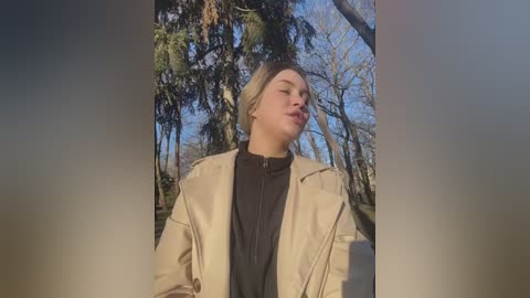 Media: A video of a young, light-skinned woman with short, light brown hair and a beige jacket, standing in a forest with tall trees and a clear blue sky.