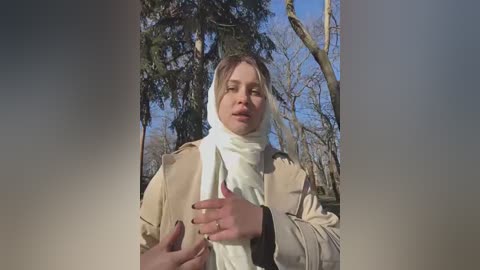 Media: A video of a Caucasian woman with light brown hair, wearing a beige coat and white scarf, standing in a forest with trees and blue sky background.