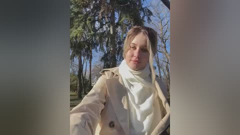 Media: Video of a blonde woman in a beige coat and white scarf, standing in a sunlit park with tall trees and a blue sky.