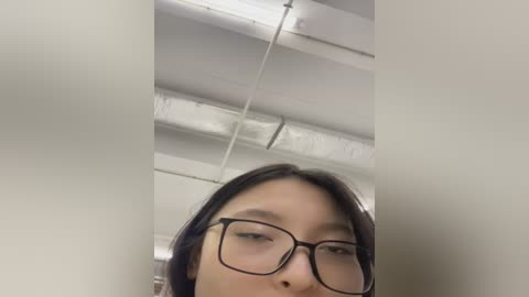 Media: Video of an Asian woman with fair skin and dark hair wearing black-framed glasses, taken from a low angle, showing her face and upper body in an indoor setting with white ceiling and exposed ductwork.