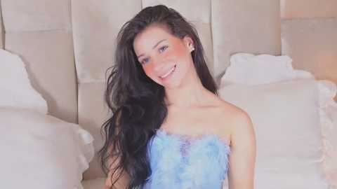 Media: Video of a smiling, light-skinned woman with long, wavy black hair, wearing a strapless, blue feathered dress, sitting on a white bed with ruffled pillows.