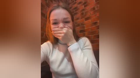 Media: A video of a young woman with straight, light brown hair, wearing a white ribbed sweater, covering her mouth with her hand. She stands against a red brick wall in a dimly lit room.