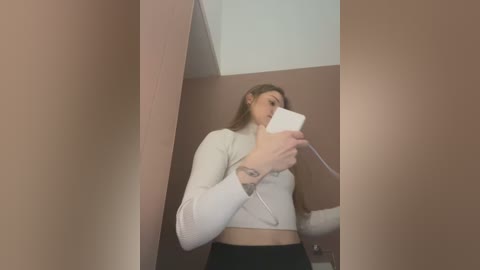 Media: Video of a young woman with long blonde hair, wearing a white crop top and black pants, taking a selfie in a dimly lit bathroom with a beige wall and white ceiling.