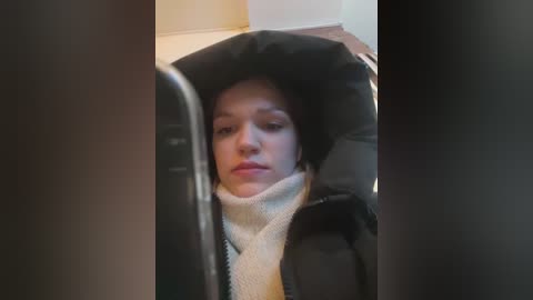 A video of a young woman in a black car seat, wearing a beige knitted scarf and a black jacket, with a neutral expression, taken indoors.