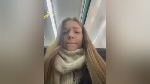 Video of a young girl with long blonde hair, wearing a cream-colored scarf and knit sweater, sitting in a train compartment with a gray seat and yellow overhead light.