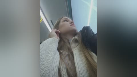 Video of a young woman with long blonde hair, wearing a beige knitted cardigan, leaning against a window in a dimly lit train, looking upward thoughtfully.