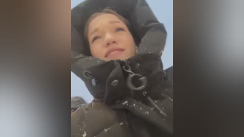 Video of a young Asian woman with light skin, wearing a black puffer jacket, snowflakes on her hood, smiling, and holding her arms up, taken from a low angle.