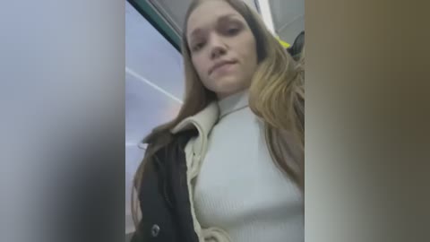 Media: A video of a young woman with fair skin, long brown hair, wearing a white turtleneck sweater and a dark jacket, looking down in a public restroom.