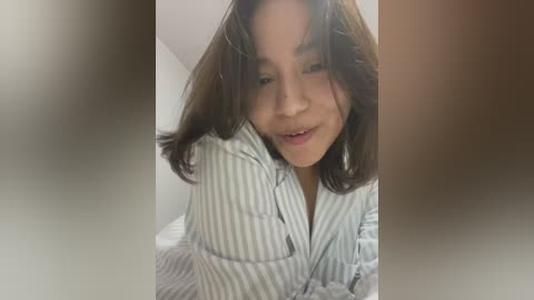 Media: A video of a young woman with shoulder-length dark hair, wearing a striped pajama top, lying on a bed. The background is blurred with soft lighting, giving a cozy, intimate feel.