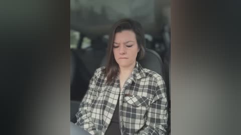 Video of a woman with shoulder-length brown hair, wearing a black shirt and a black-and-white plaid flannel, seated in a car with a blurred, indistinct background.