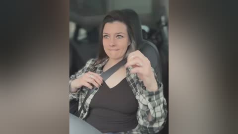 Media: Video of a woman in a car, wearing a black top and plaid shirt, adjusting her seatbelt. The background is blurred, showing a car interior.