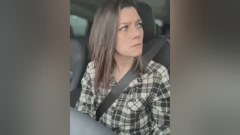 Media: Video of a young woman with straight brown hair, wearing a black and white plaid shirt, seated in a car with a seatbelt on, looking contemplative and slightly sad.