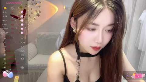 Media: A video of an East Asian woman with long black hair, fair skin, and red lipstick, wearing a black choker and a black top, in a modern, minimalist room.