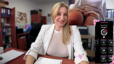 Media: Video of a smiling blonde woman in a white blazer over a striped shirt, sitting at a desk in an office with bookshelves and computer screens.