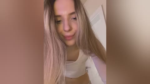 Media: Video of a young woman with long, straight, blonde hair, wearing a white crop top, looking downward with a slight smile. Background shows a blurred white door.