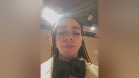 Media: A video of a young woman with long brown hair wearing a white shirt and black headphones, taken indoors with a soft, warm lighting.