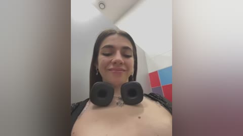 Media: Video of a young woman with light skin, long brown hair, and black headphones covering her small breasts in a minimalist, brightly lit room.