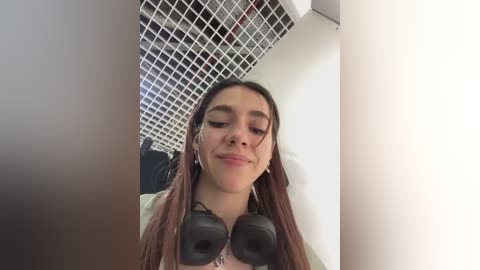 Media: Video of a young woman with long brown hair, wearing black headphones, smiling in a dimly lit room with white walls and a grid-patterned ceiling.