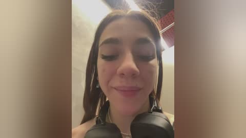 Media: Video of a young woman with light skin and long brown hair, wearing black headphones, smiling softly, with closed eyes, in a dimly lit room with red and beige walls.