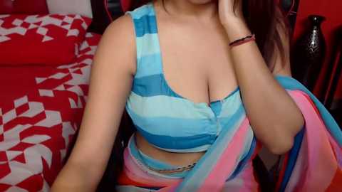 Media: Video of a young woman with light brown skin, wearing a blue and white striped crop top that accentuates her large breasts. She sits on a bed with a red and white patterned duvet, her hand on her face, revealing a bracelet.