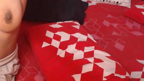 Media: Video of a nude woman lying on a red bedspread with a white geometric pattern, wearing a beige shirt and black pillow, showing her left breast and nipple.