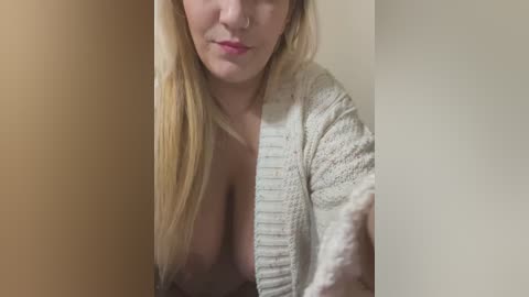 Media: A video of a blonde woman with fair skin, wearing a white knitted cardigan, exposing her large breasts, partially obscured by a shadow. The background is a beige wall, creating a stark contrast with her light hair and skin tone.