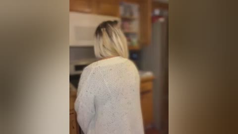 Media: Video of a blonde woman with short hair, wearing a light-colored, knitted sweater, standing in a dimly lit kitchen with wooden cabinets and a stainless steel fridge in the background.