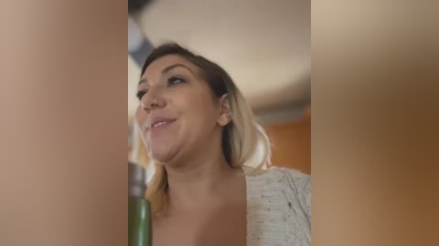 Media: A close-up video of a young woman with light brown skin, light brown hair, and a white top, smiling while drinking from a green bottle. The background is blurred, featuring warm-toned furniture.